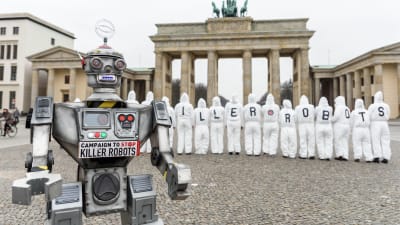 En robot i förgrunden och aktivister i bakgrunden som demonstrerar mot mördarrobotar framför Brandenburger Tor i Berlin.