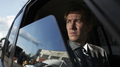 Pasi tittar ut genomen ett öppet bilfönster som speglar en fabrik. 