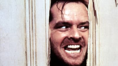 Närbild på Jack Torrance (Jack Nicholson) då han med yxa huggit ett hål i en dörr och nu med ett galet leende tittar in genom hålet.