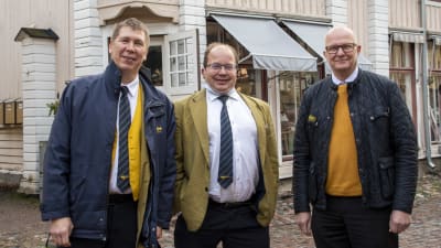 Tre manliga busschaufförer står i ett gammalt trähuskvarter och tittar in i kameran och ler.