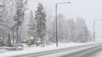 En grävmaskin arbetar vid sidan av en riksväg. Det snöar ymnigt.