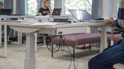 En lärare i ett klassrum sitter vid ett höj- och sänkbart bord med sin laptop.