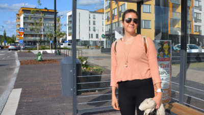En kvinna i ljus tröja och solglasögon står vid en busshållplats i Nickby.