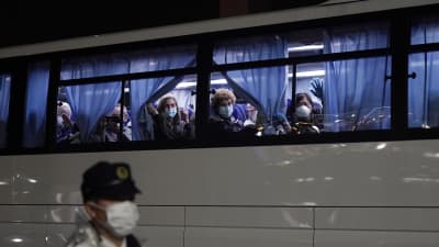 Amerikanska medborgare sitter i en buss och tittar ut ut fönstret. Bussen har blå gardiner som passagerarna håller i. Utanför bussen syns en person med ett andningsskydd och vit rock.