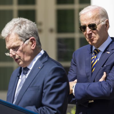 Joe Biden i förgrunden som håller armarna i kors. Han har på sig en kostym och pilotsolglasögon. I bakgrunden Sauli Niinistö i kostym vid ett talarpodium.