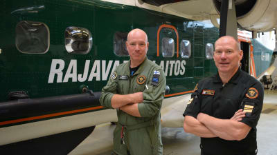 Två män med väldigt kort hår står med armarna i kors framför Gränsbevakningsväsendets gröna flygplan. Den ena mannen har skogsgrön overall, den andra mörkblå tjänsteskjorta.