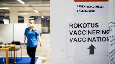 Skylt bland annat med texten vaccinering och till vänster en kvinna som bär munskydd. 85-åringar och äldre samt närvårdare vaccinerades på Busholmen i Helsingfors den 1 februari  2021.