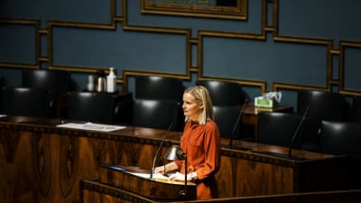 Riikka Purra i orange klänning vid plenisalens podium