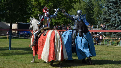 Två riddare i rustning till häst ger varandra en high five. Den ena hästen har rödvitt täcke, den andra blått.