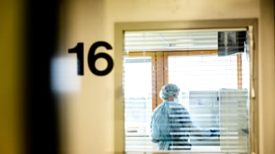En sjukskötare jobbar i isoleringsrum för coronapatienter, fotograferad genom glasdörr.