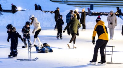 Både barn och vuxna som åker skridskor på en skridskoplan med snövallar runt omkring.