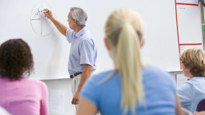 En äldre manlig lärare skriver på en whiteboard i ett klassrum med elever i förgrunden av bilden.