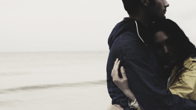 Kvinna i gul jacka kramar en längre man på en strand i dimmigt väder.