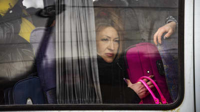 En kvinna sitter i en buss med en väska i famnen. Hon är fotograferad från fönstret.