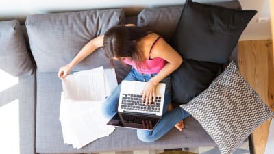 En kvinna sitter i en soffa med sin dator och många papper. Bilden är tagen uppifrån.