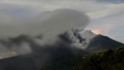 Ett tre kilometer högt och gråsvart askmoln stiger från vulkanen Turrialba i Costa Rica och rör sig mot huvuddstaden San José vars ljus syns i bakgrunden.