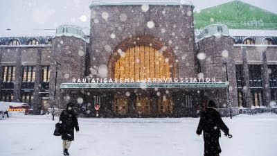 Två personer utanför järnvägsstationen i Helsingfors. Det snöar.