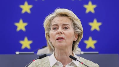Ursula von der Leyen står och talar. Hon tittar framåt, något åt sidan. I bakgrunden EU:s flagga på en vägg.