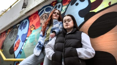 Kaksi nuorta naista nojaavat graffitiseinää vasten.