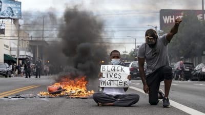 En man sitter på marken bredvis något som brinner. Han håller upp skylt där det står att svarta liv är värda lika mycket. Bredvid står en annan man på huk, han håller upp handen.