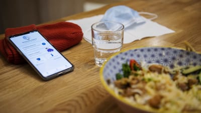 En mobil med Coronablinkern på skärmen ligger lutad mot en mössa på ett bord. Framför den en portion med nudlar, ett vattenglas och ett munskydd.