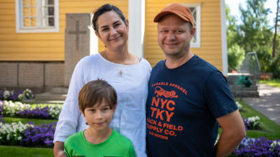 En familj, mamma i vit skjorta och mörkbrunt hår, pappa i orange keps och blå t-skjorta och liten son i grön t-skjorta och mörkt hår.