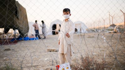 En liten pojke i munskydd tittar in i kameran genom stängsel på gränsen mellan Afghanistan och Iran. I bakgrunden syns tält och två män som pratar med varandra.