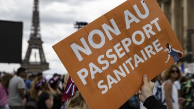 Bild på en orange skylt där det står "nej till hälsopass" skrivet på franska. Bakom syns en folkmassa och Eiffeltornet.