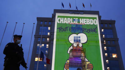 Bild på polis med munskydd utanför byggnad där en stor bild av satirtidningen Charlie Hebdos uppslag projiceras på fasaden till minne av Samuel Paty.