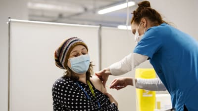 Kvinna iklädd i munskydd får en spruta i armen av en sköterska.