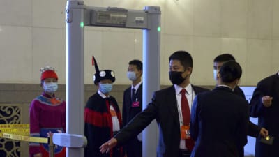 Folk går igenom en säkerhetskontroll för att komma in på folkkongressen i Kina. I förgrunden en man som iklädd andningsskydd och kostym går igenom en metalldetektor.