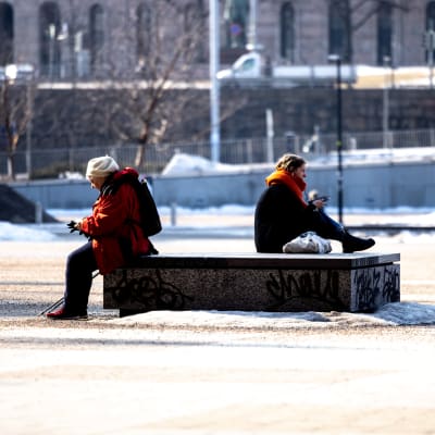 En äldre kvinna och en yngre man sitter med ryggarna mot varandra vid en staty. 