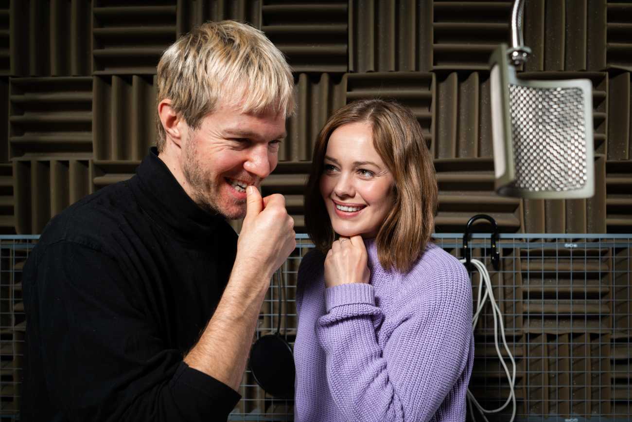 Kuvassa hymyilev mies- ja naisnyttelij seisovat kuunnelmastudiossa katosta roikkuvan mikrofonin vieress.