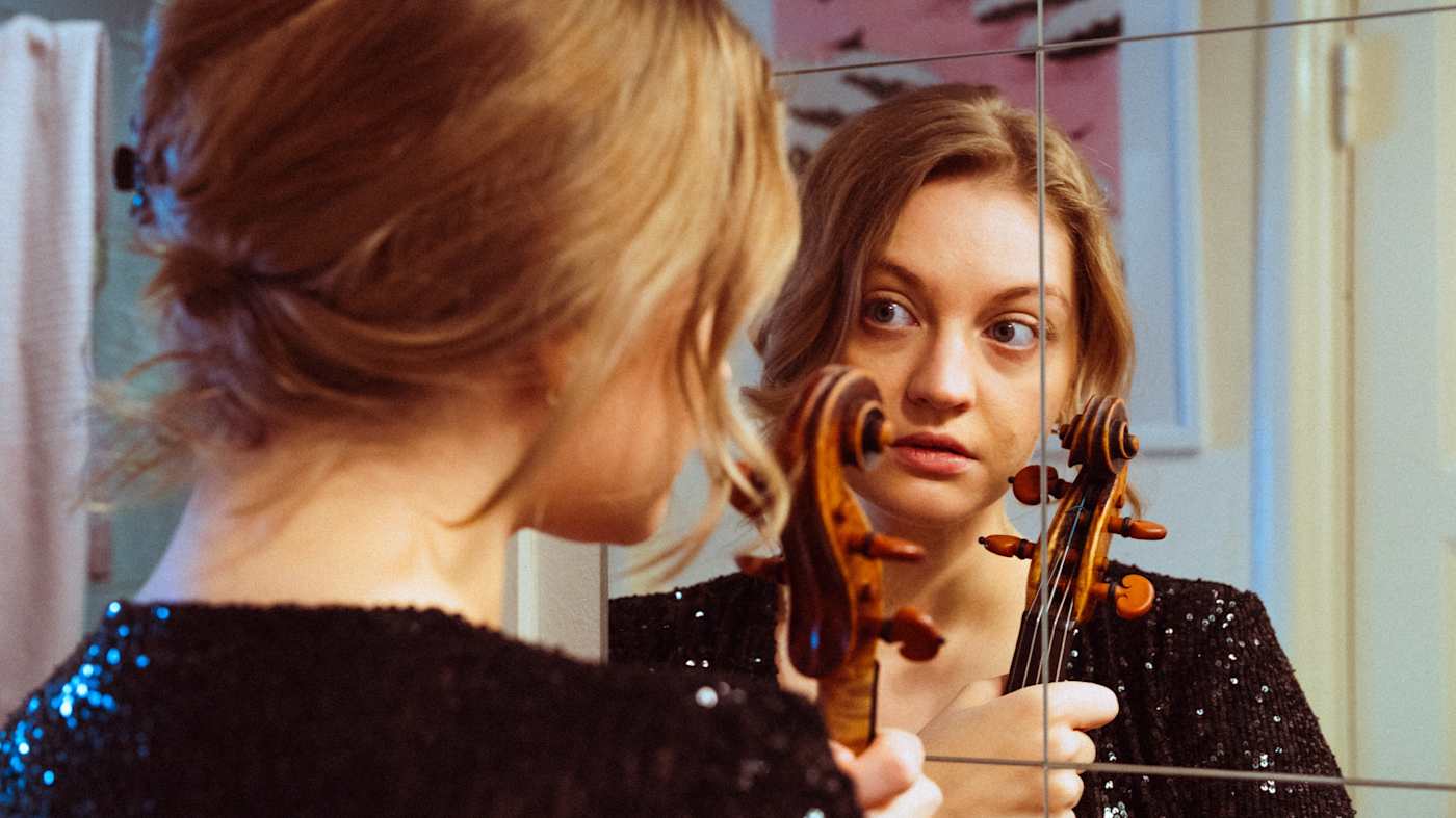 Nuori nainen seisoo peilin edessä, hän pitelee viulua.