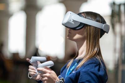 En blond kvinna står med VR-glasögon på sig och håller i två styrhandtag.