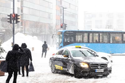 Jalankulkijoita, taksi ja bussi lumisateessa.