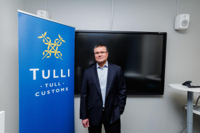 Tullens övervakningsdirektör Sami Rakshit står bredvid en blå skärm där det står Tulli, tull, customs. 