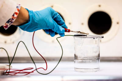 En hand i blå plasthandske håller en liten kabel i ett vattenglas för att genomföra elektrolys.