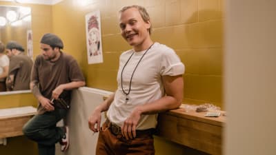 Nuori mies (näyttelijä Elias Salonen) valkoisessa t-paidassa ja ruskeissa dongareissa nojailee hymyillen kylpyhuoneen kaakeloidulla seinällä olevaan kapeaan hyllyyn.