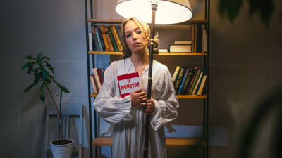 Nuori nainen (näyttelijä Aamu Milonoff) katsoo kameraan pidellen toisessa kädessään kirjaa "La Monstre" ja toisessa jalkalamppua, taustalla kirjahylly sekä viherkasvi tuolin päällä.