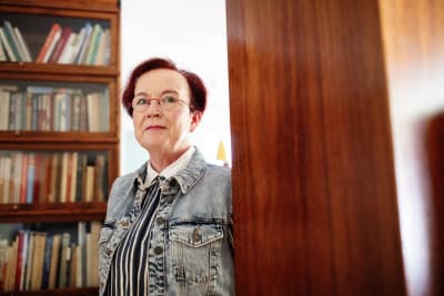 Seija Ilmakunnas ser in i kameran bakom en hylla, i bakgrunden en bokhylla.