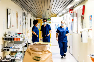 Tre sjukskötare med blå arbetskläder och munskydd går i en sjukhuskorridor.