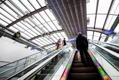 En rulltrappa som för tittaren uppåt mot en tågstations hall. Taket är gjort av glas och det hänger informationsskyltar i bakgrunden.