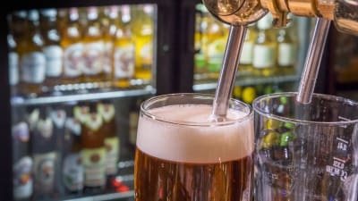 Två ölglas, med öl i ett av dem. I bakgrunden syns ölflaskor i kylskåp.