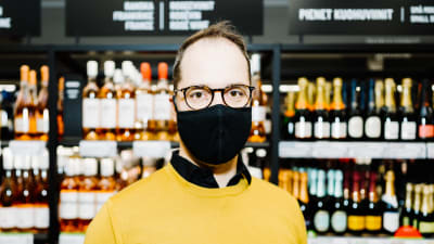 En man klädd i gul tröja står framför en butikshylla med drycker på i Alko. Han har munskydd på sig och tittar in i kameran.