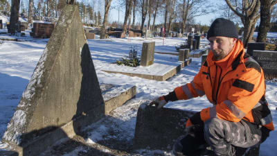 En man i orange arbetskläder sitter på huk och pekar på en gravsten.