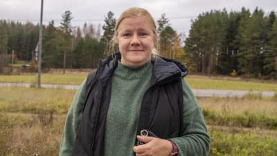 En kvinna står utomhus och tittar in i kameran. I bakgrunden syns en väg och skog.