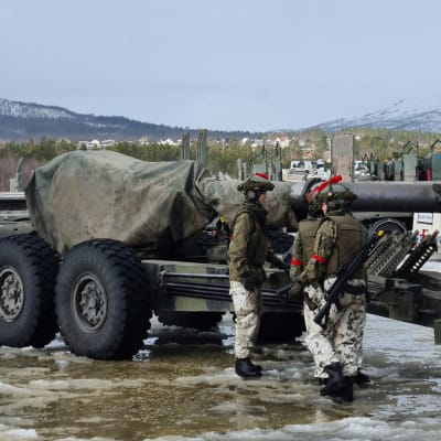 Några värnpliktiga står vid en övertäckt artilleripjäs i ett vårtöigt fjällanskap. Deras camouflagedräkter är i huvudsak vita nedtill och grönspräckliga på överkroppen.