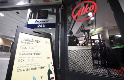 Till vänster syns en skylt där öppettider står listade, till vänster en skylt med texten alko. I bakgrunden syns en person med en kundvagn.