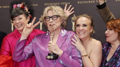 Fyra festklädda kvinnor ser glada ut. En av dem håller en statyett i ena handen och visar segertecken med den andra. 